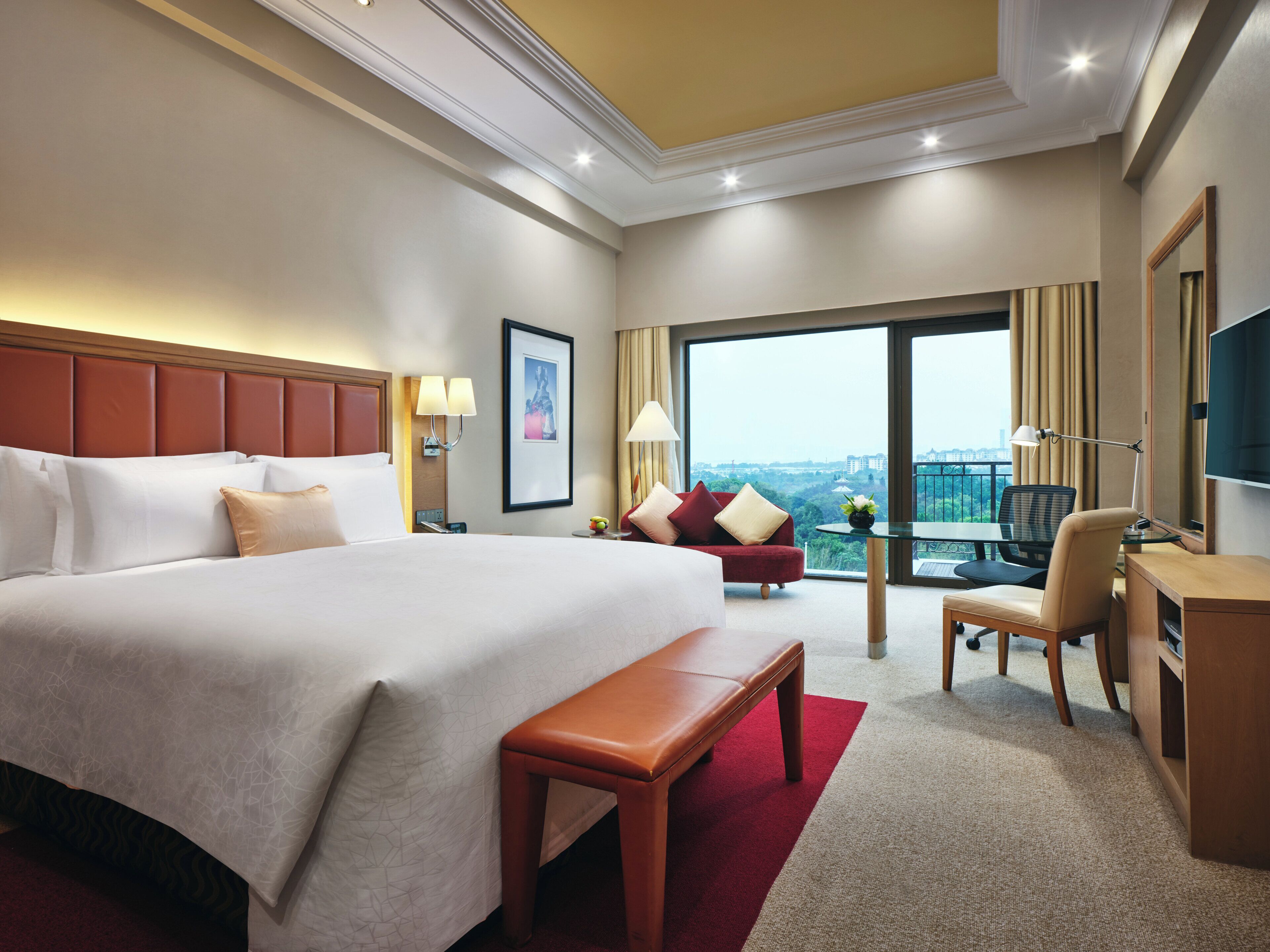 A hotel room with a large bed, a desk, a chair, a small table, a bench, and a television.