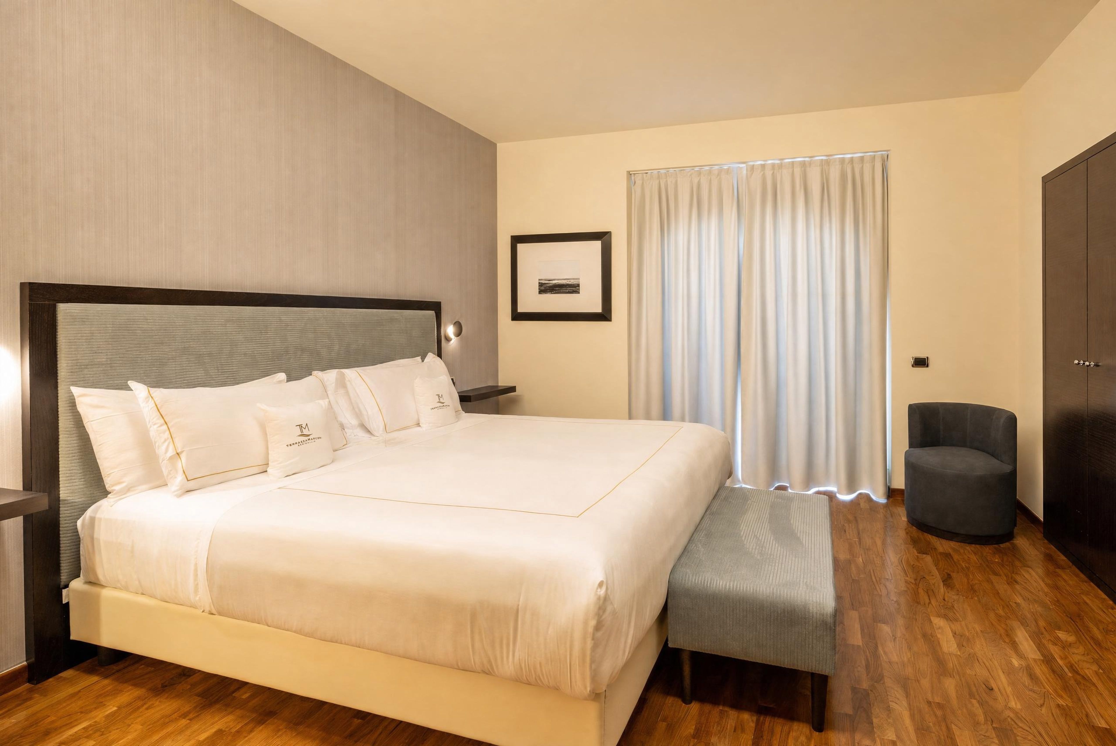 Une chambre d’hôtel avec un grand lit, une table de chevet, une chaise, une armoire et une fenêtre avec des rideaux.