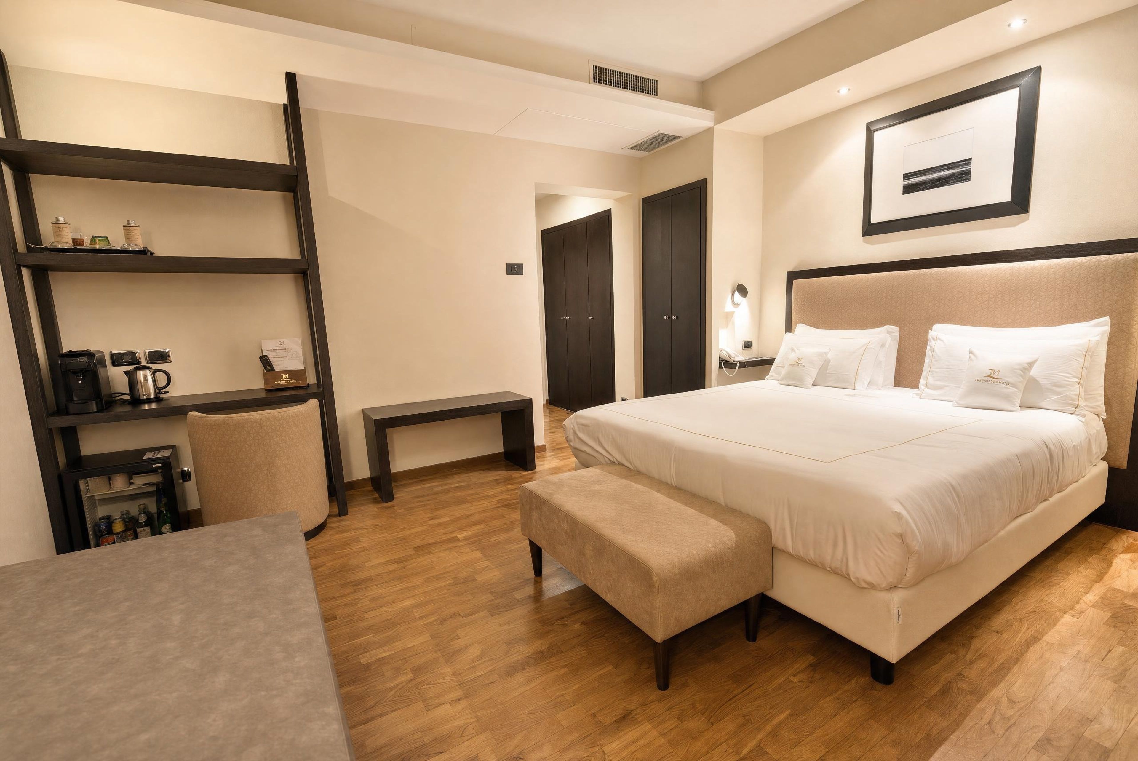 A hotel room with a large bed, a desk, a chair, a bench, and a shelf with items.