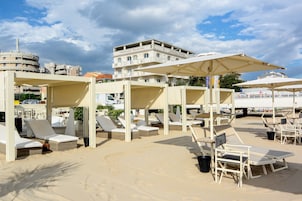 Un área de descanso frente a la playa con tumbonas y sombrillas, una playa arenosa y un edificio al fondo.