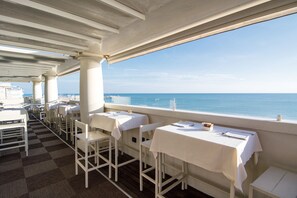 Een restaurant met witte tafels en stoelen, een geruit vloertje en uitzicht op zee.