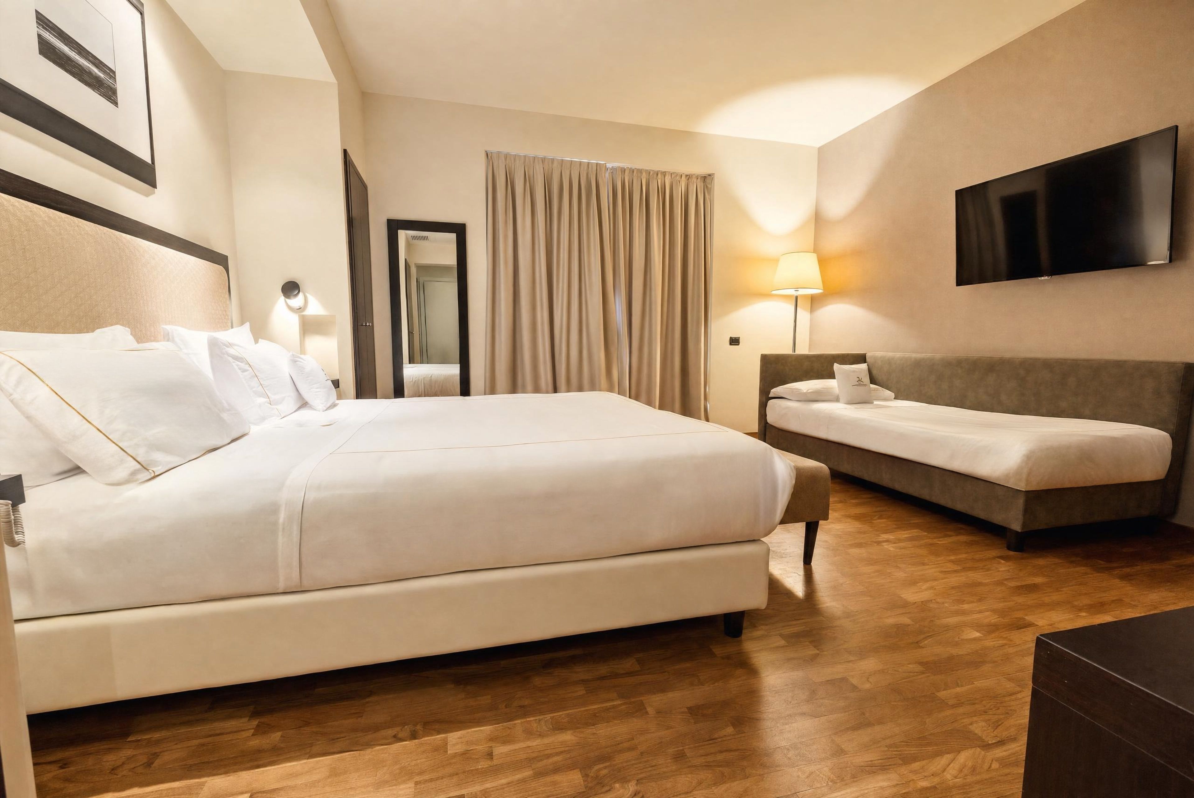 A hotel room with a large bed, a sofa, a flat-screen TV, and a mirror.