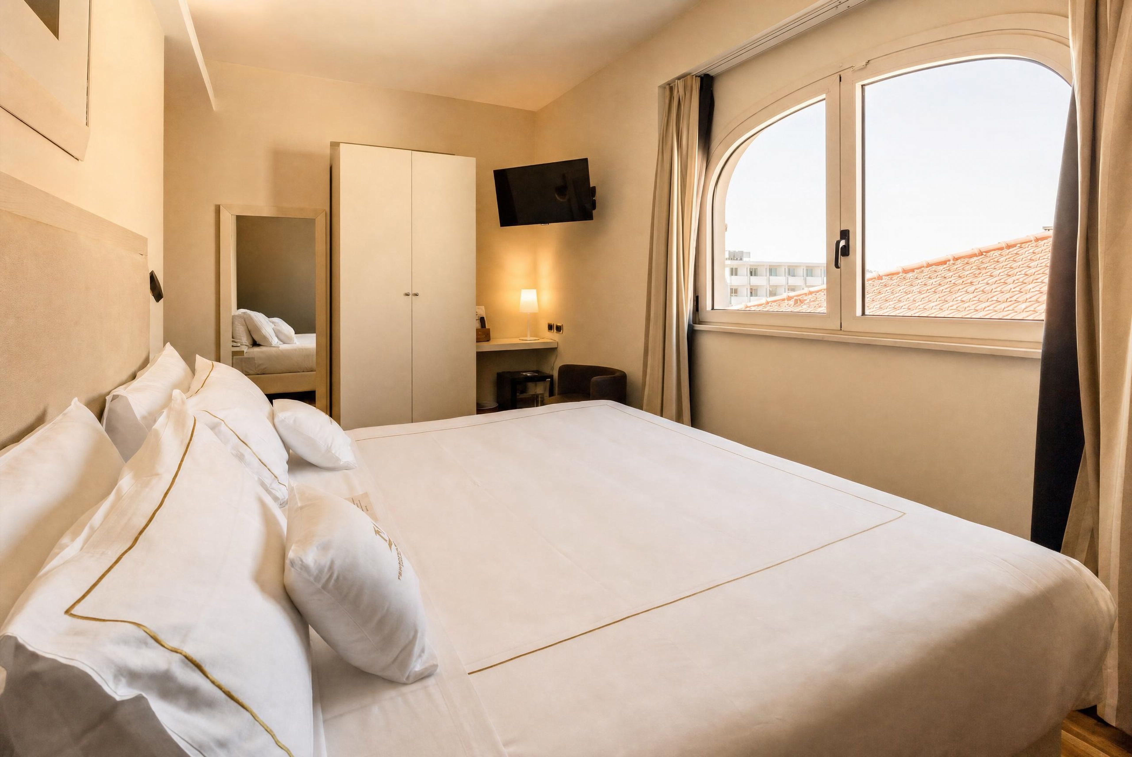 A hotel room with a large bed, a wardrobe, a TV mounted on the wall, and a window with curtains.