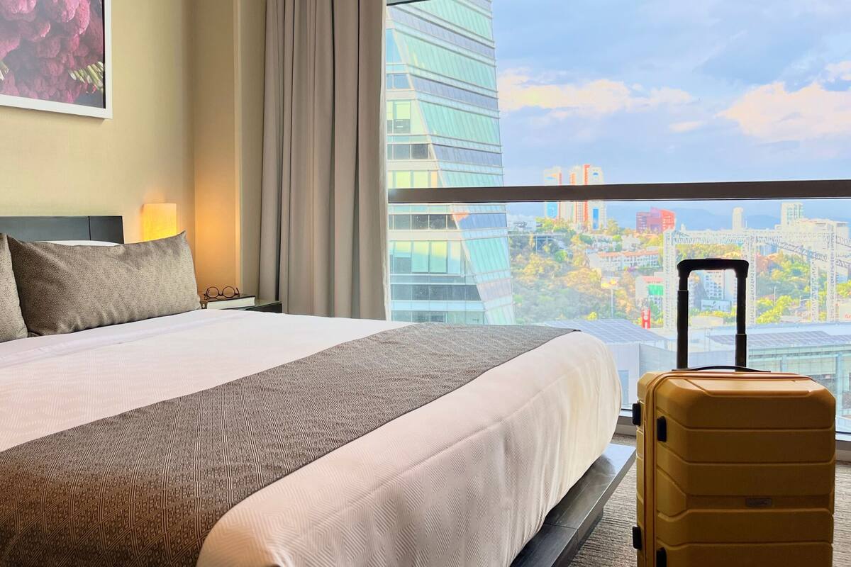 A hotel room with a large bed, a view of the city, and a yellow suitcase.