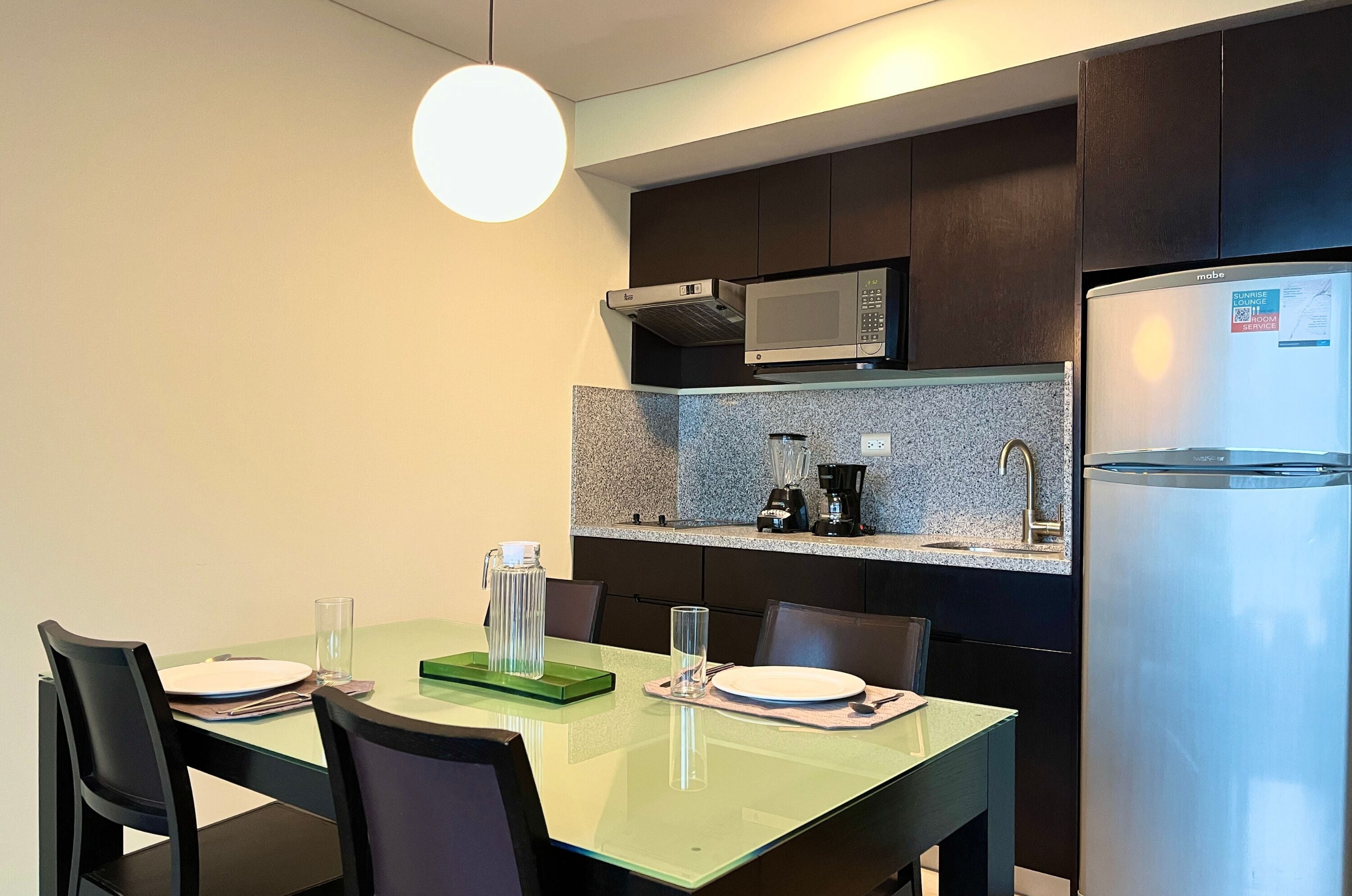 A modern kitchen with a microwave, coffee maker, and refrigerator. There is a dining table with chairs and a glass top.