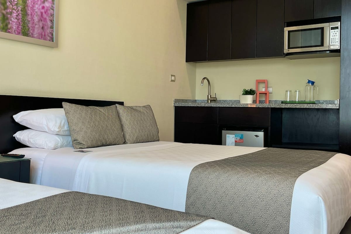 A hotel room with two beds, a nightstand, a lamp, a microwave, a small kitchen area, and a wall-mounted shelf.