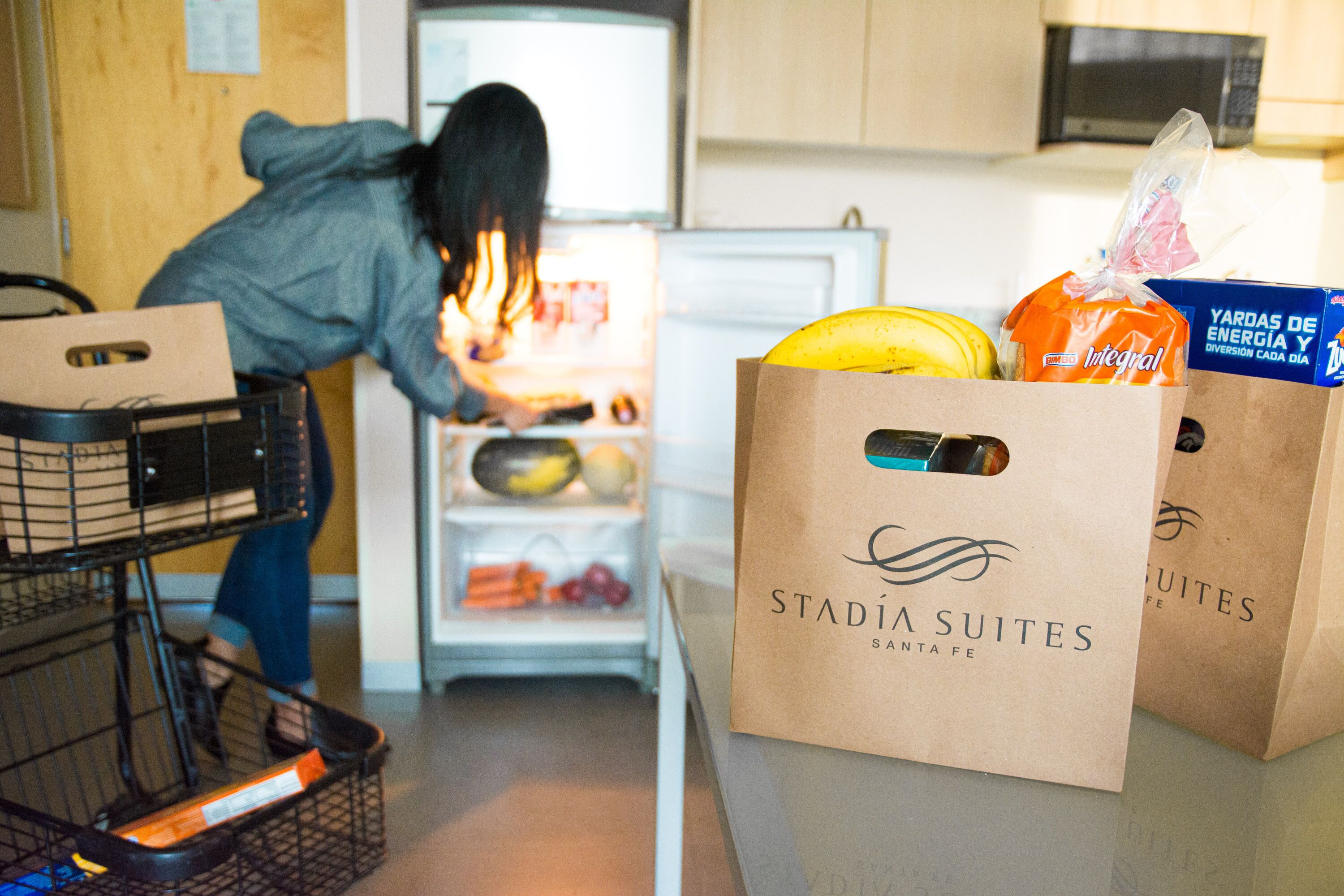 Una cocina con un refrigerador lleno de comida, una cesta de compras y dos bolsas de papel de 'Stadia Suites'.