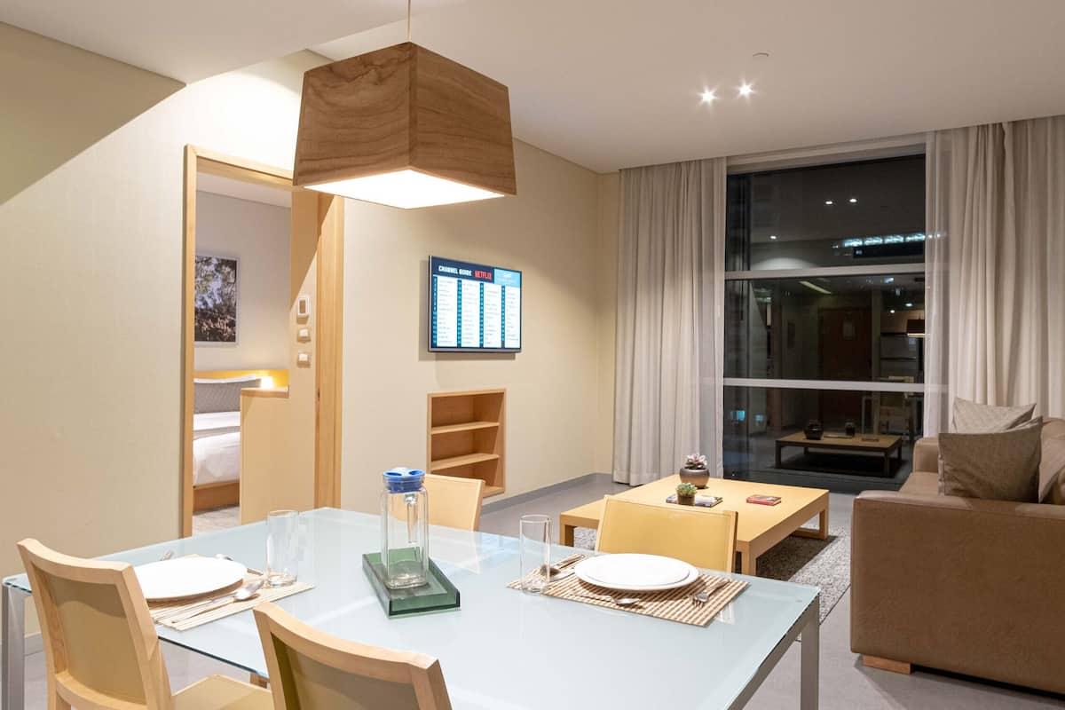 A modern hotel room with a dining area, a sofa, a TV displaying a channel list, and a large window with curtains.