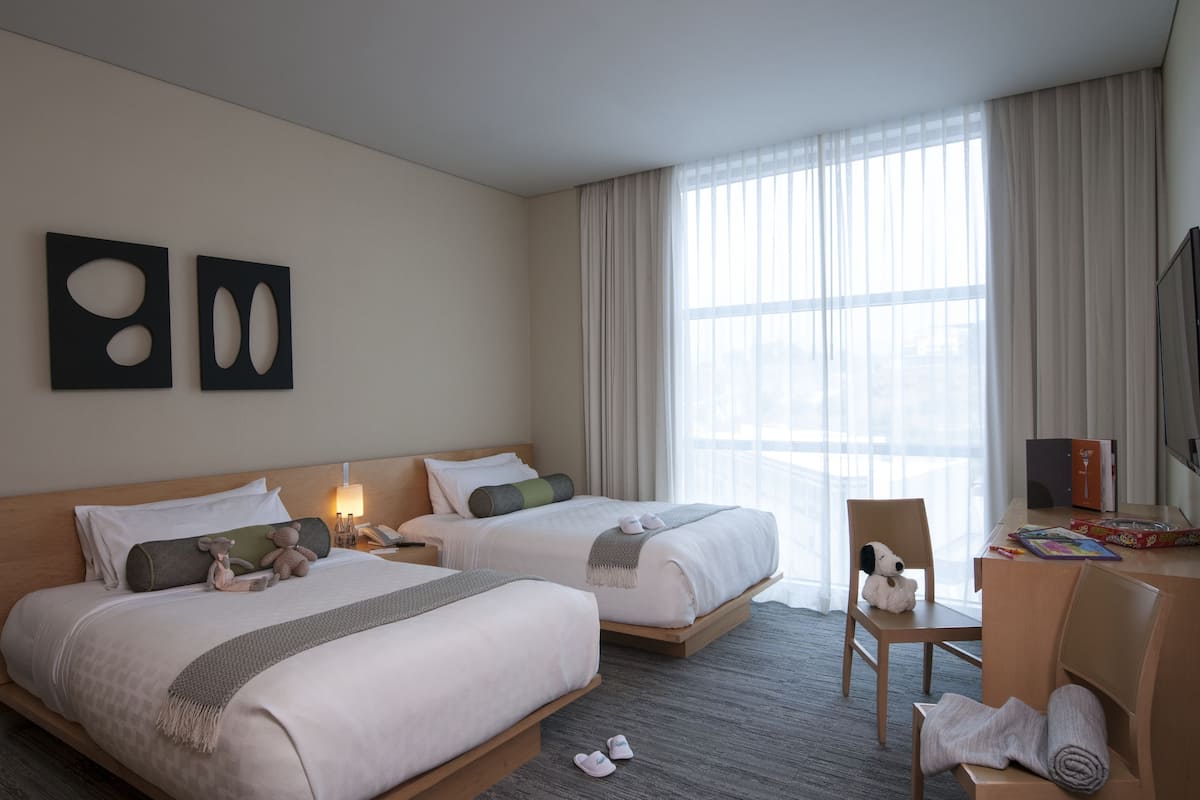 A hotel room with two beds, a desk, a chair, and a large window with curtains.