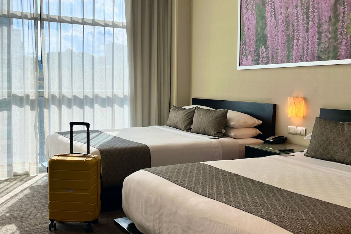 A hotel room with two beds, a bedside table with a lamp, a suitcase, and a large window with sheer curtains.