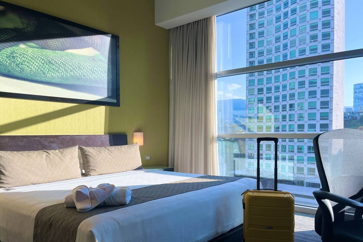 A hotel room with a bed, a suitcase, a chair, and a large window with a city view.