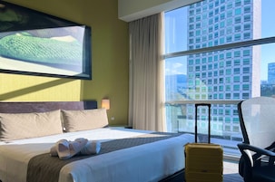 Habitación de hotel con cama, maleta, silla y ventana grande con vista a la ciudad.