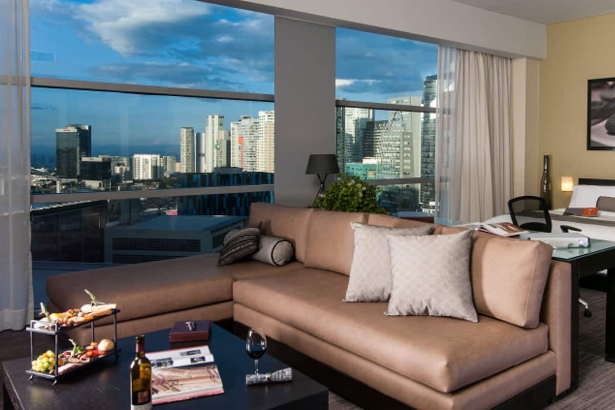 A modern hotel room with a large window offering a city view, a sofa, a bed, and a coffee table with amenities.