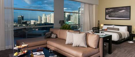 A modern hotel room with a large window offering a city view, a sofa, a bed, and a coffee table with amenities.
