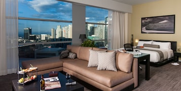 Habitación de hotel moderna con un amplio ventanal que ofrece vistas a la ciudad, un sofá, una cama y una mesita con servicios.