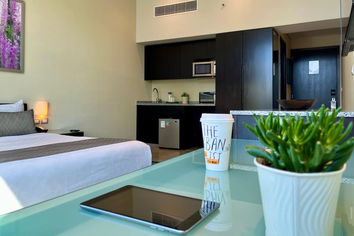 A hotel room with a bed, a bedside table with a tablet, a coffee cup, a potted plant, a kitchenette with a microwave, and a door leading to another room.