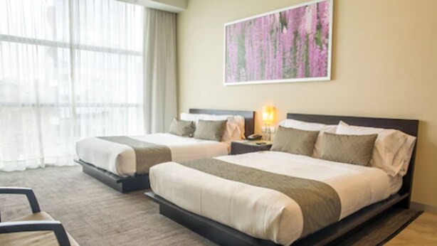 A hotel room with two beds, a nightstand with a lamp, a chair, and a large window with sheer curtains. - Stadía Suites Mexico City Santa Fe (Mexico City)