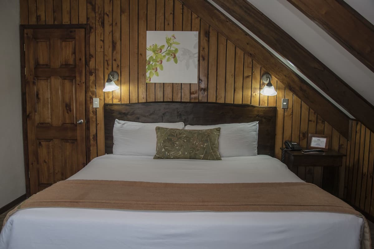 A bedroom with a wooden ceiling, a large bed with white bedding, a brown throw blanket, and two wall-mounted lamps.
