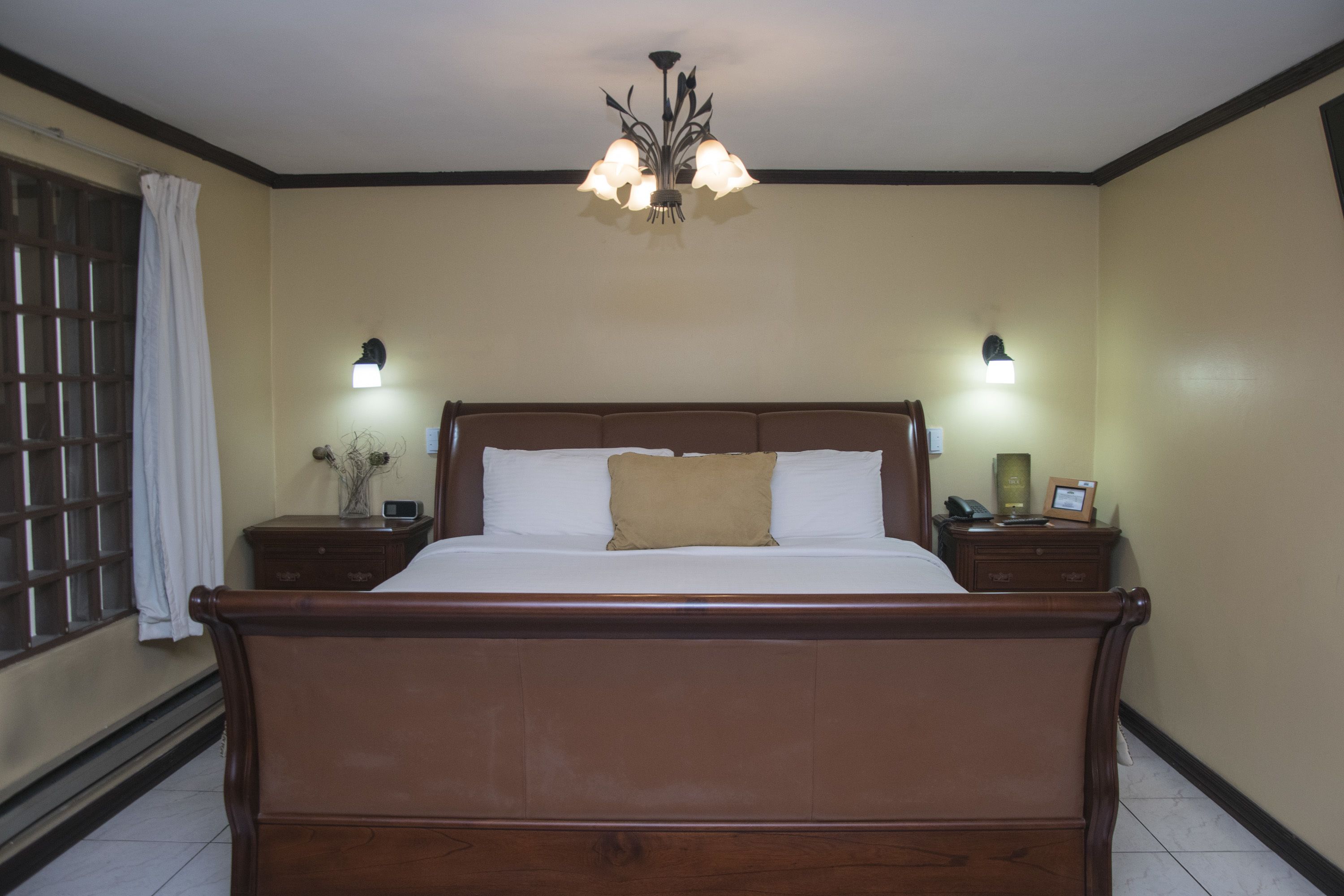 A bedroom with a wooden bed, two bedside tables, a chandelier, and a window with a curtain.