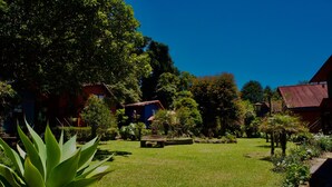 A garden with a lawn, a large tree, and a building with a red roof. - Hotel El Tirol (Ángeles)