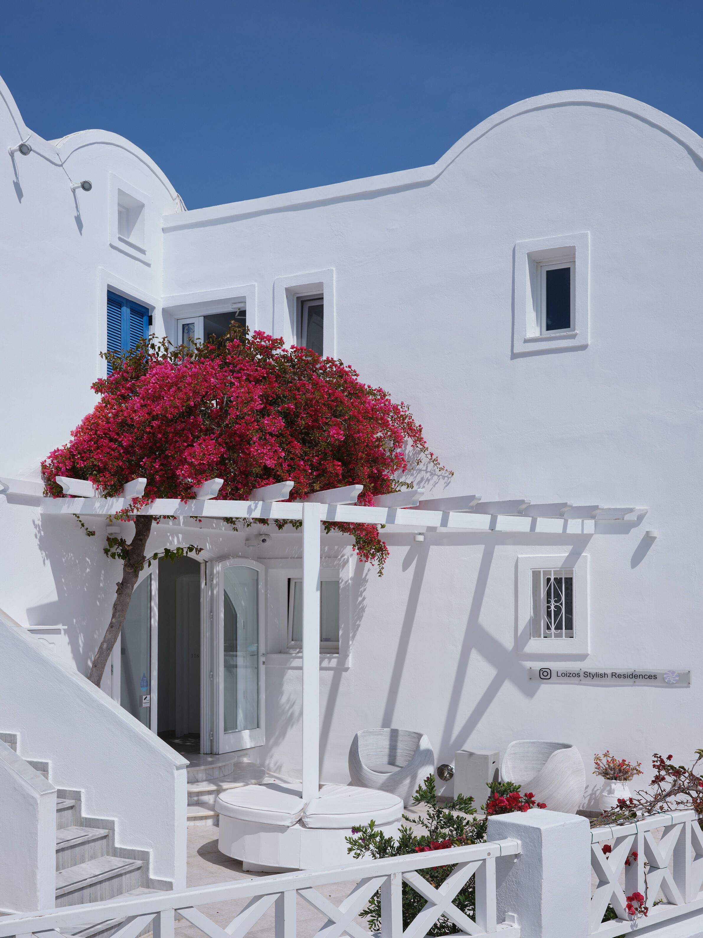 Um edifício branco com área de estar coberta ao ar livre, uma escadaria e uma pequena janela com grade metálica decorativa.