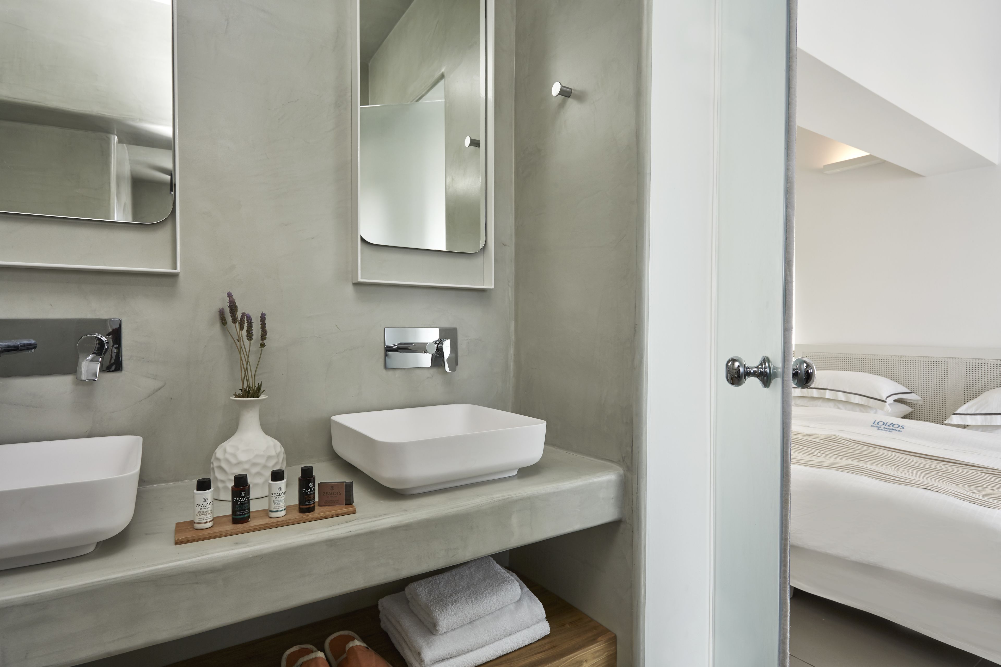 A modern bathroom with two sinks, a mirror, and a shower. There is a wooden shelf with toiletries and towels.