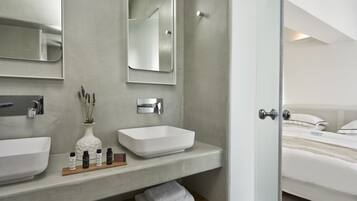 A modern bathroom with two sinks, a mirror, and a shower. There is a wooden shelf with toiletries and towels.