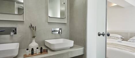A modern bathroom with two sinks, a mirror, and a shower. There is a wooden shelf with toiletries and towels.