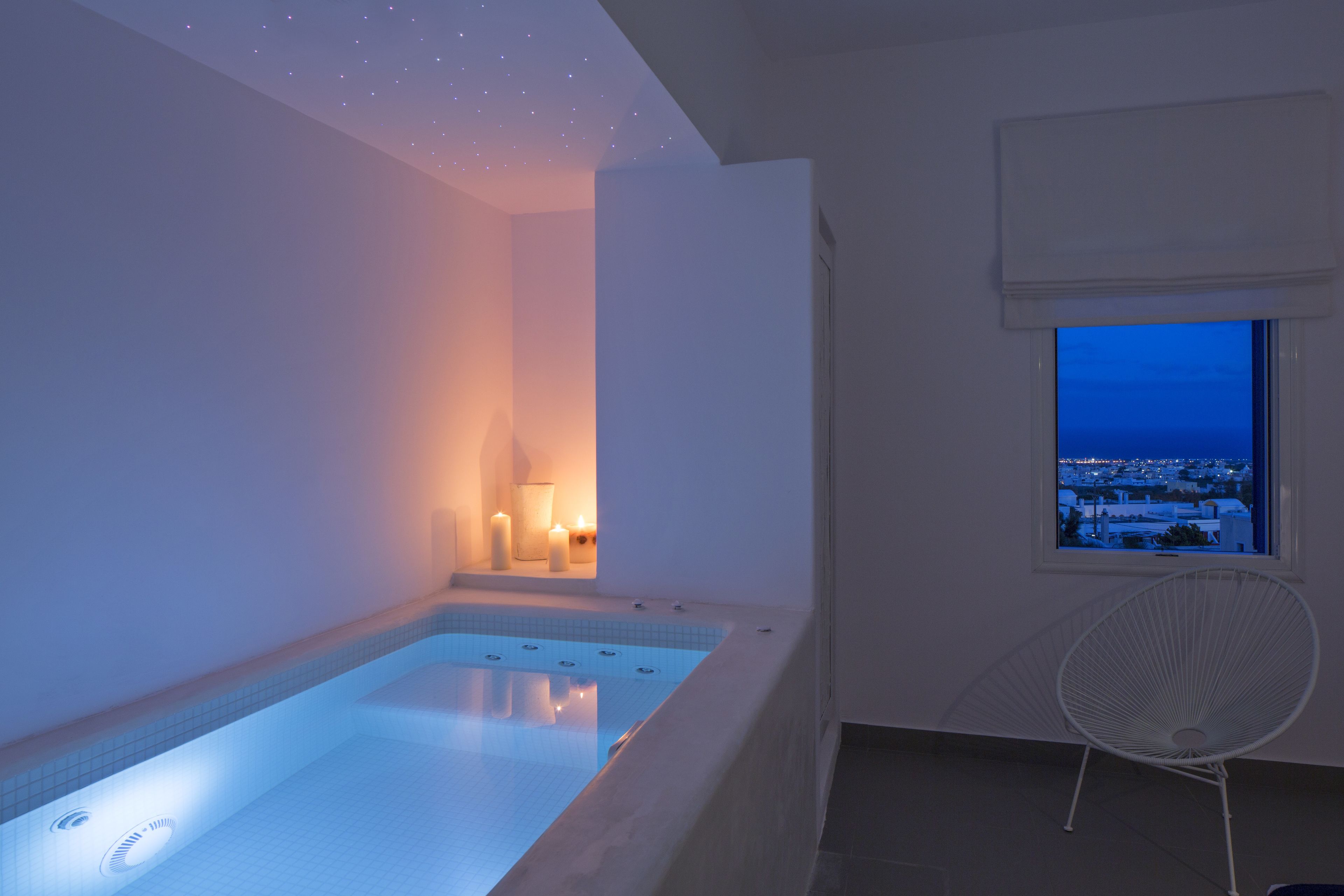 A modern indoor swimming pool with a view of a cityscape at dusk, a white chair, and a window with a view.