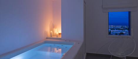 A modern indoor swimming pool with a view of a cityscape at dusk, a white chair, and a window with a view.
