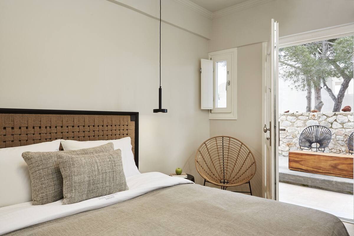 A bedroom with a bed, a wicker chair, and a stone wall visible through an open door.
