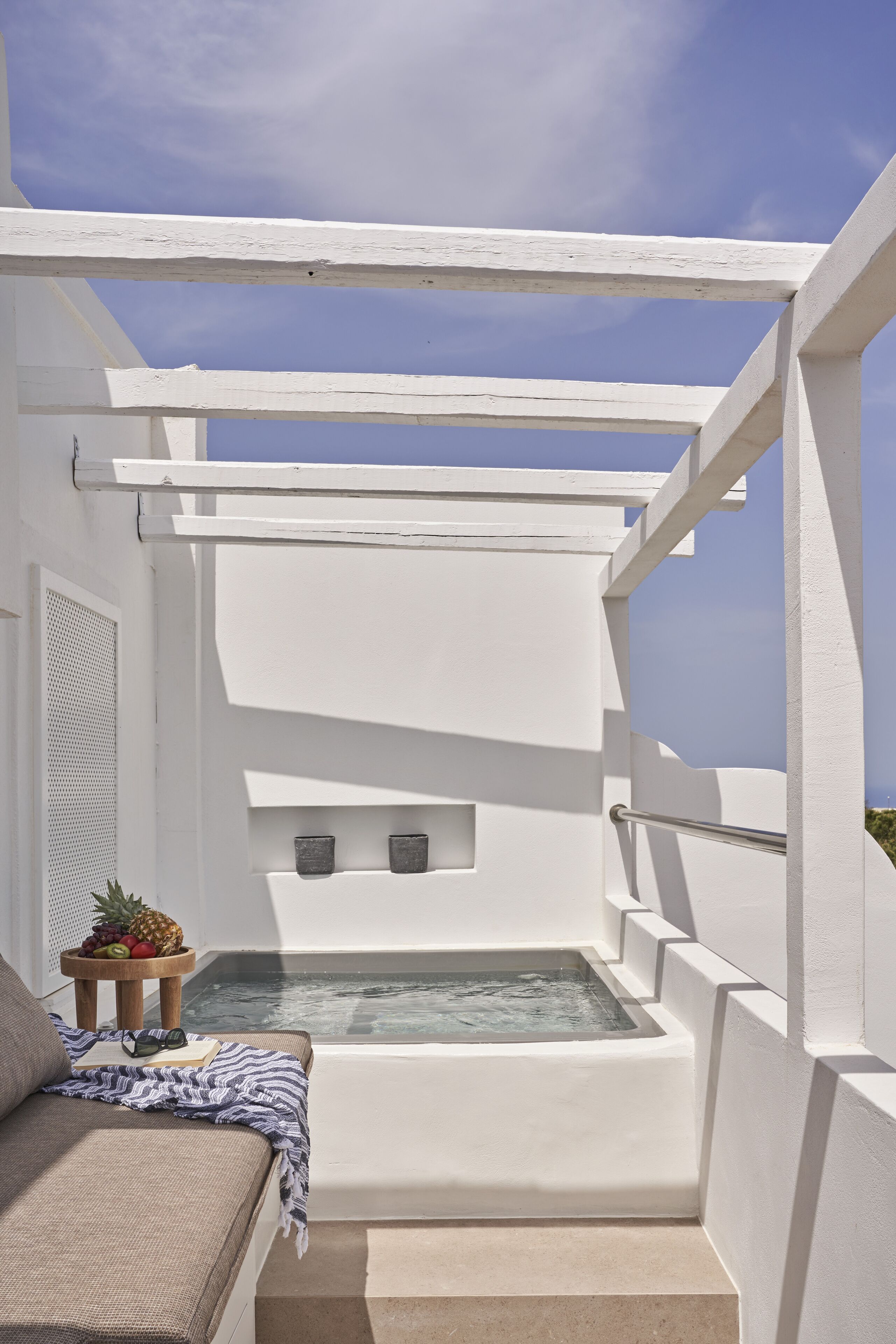A white balcony with a built-in hot tub, a cushioned seating area, and a small table with fruits and a book.