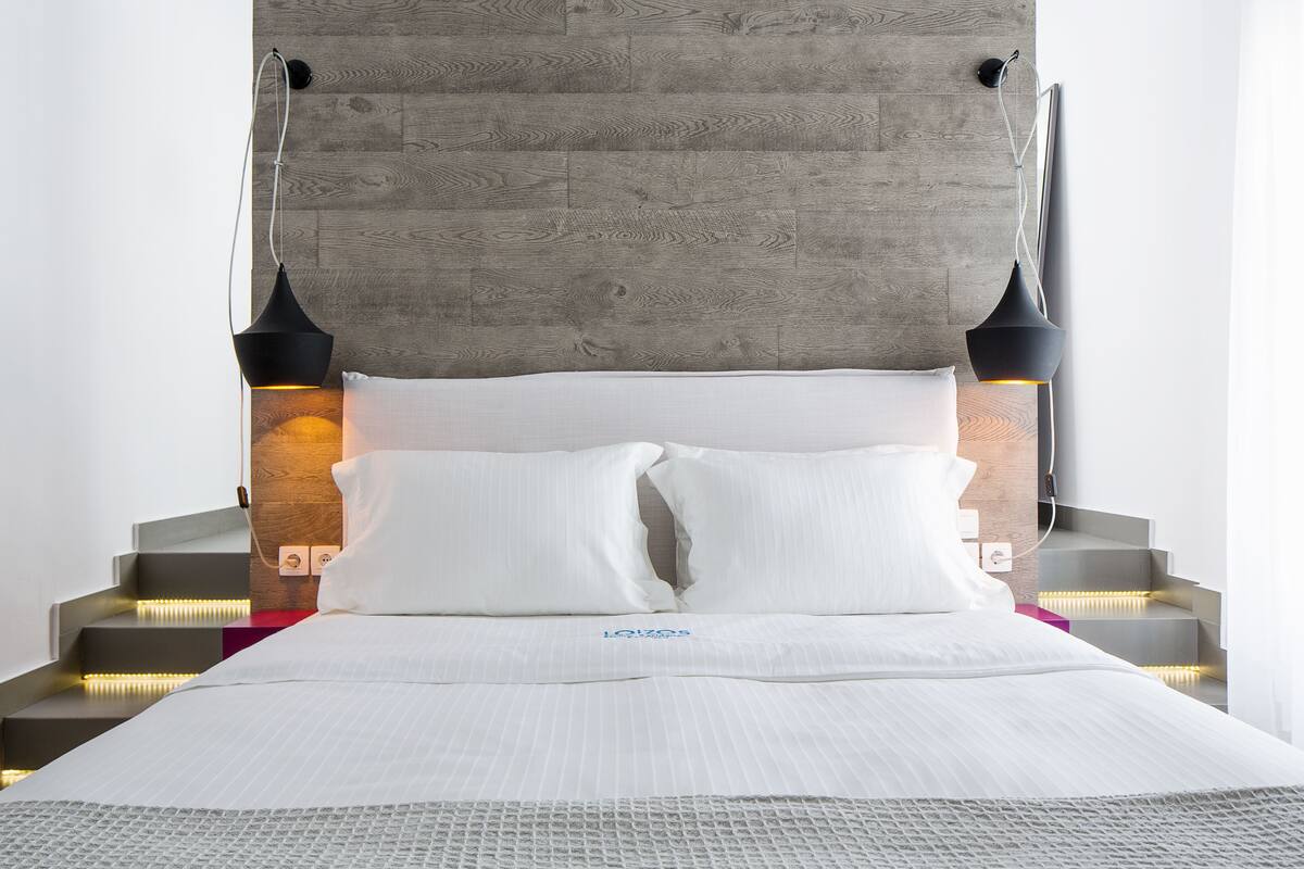 A modern hotel room with a wooden headboard, a neatly made bed with white linens, and two black pendant lights on either side.
