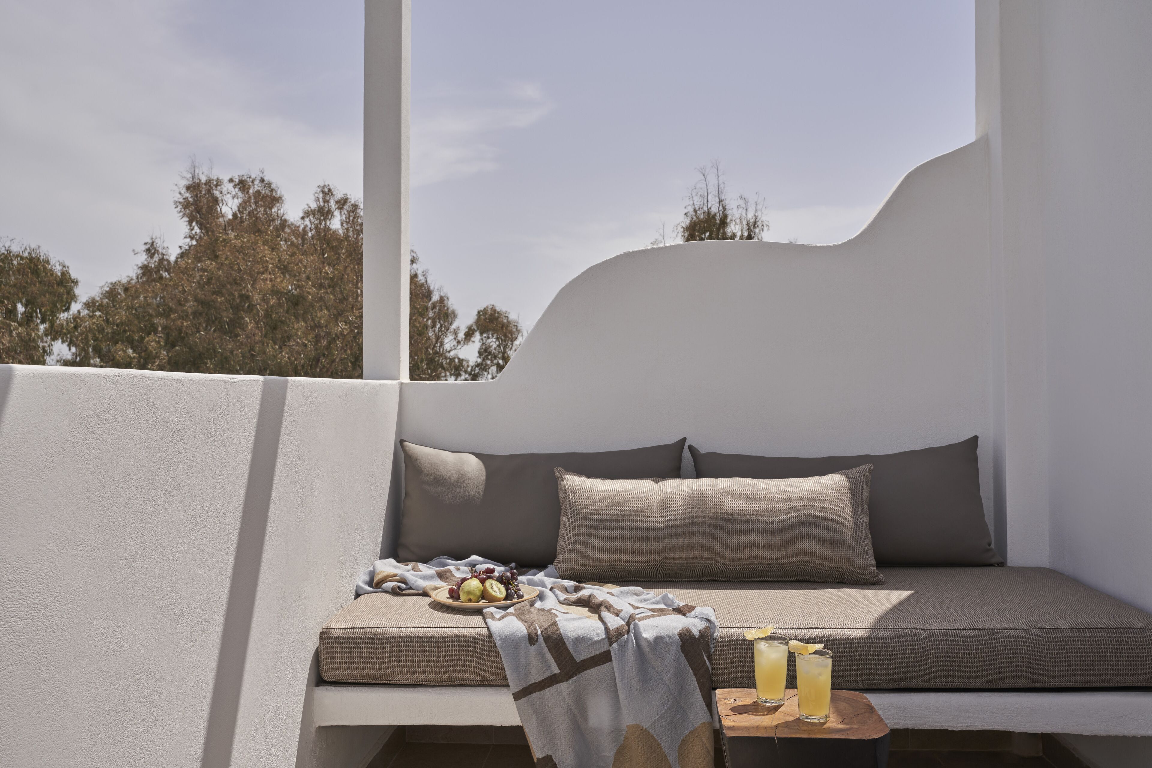 Un área de estar moderna al aire libre con un banco acolchado, una mesita con bebidas y fruta, y vistas a árboles.