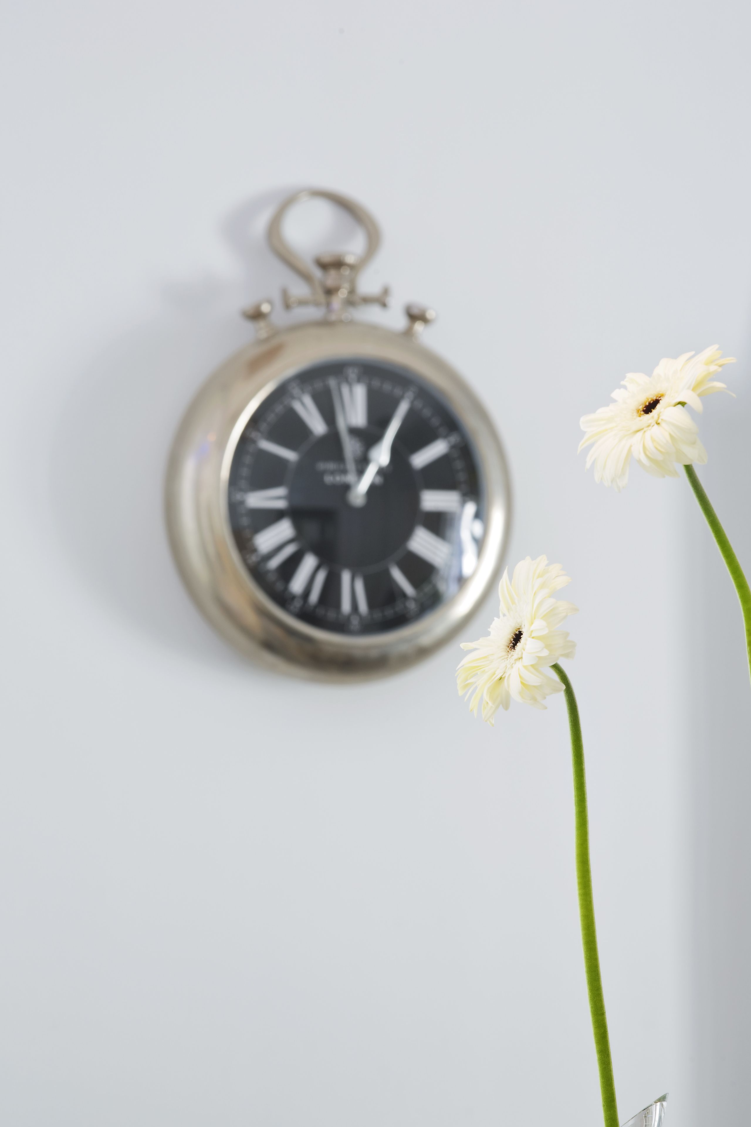Eine wandmontierte Taschenuhr mit römischen Ziffern und einer Blume vor weißem Hintergrund.