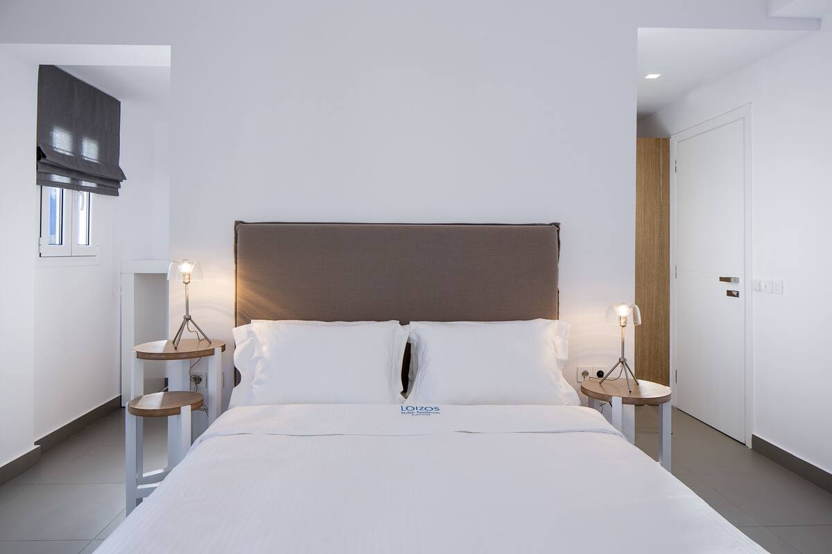 A modern hotel room with a large bed, two bedside tables with lamps, a wooden headboard, and a door to the right.