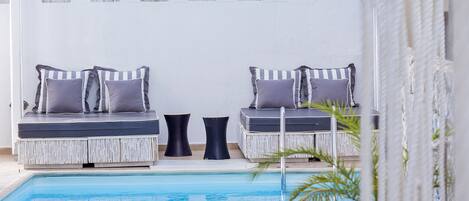 Uma área de estar à beira da piscina com dois bancos estofados e uma pequena mesa, cercada por um muro branco com janelas em arco.