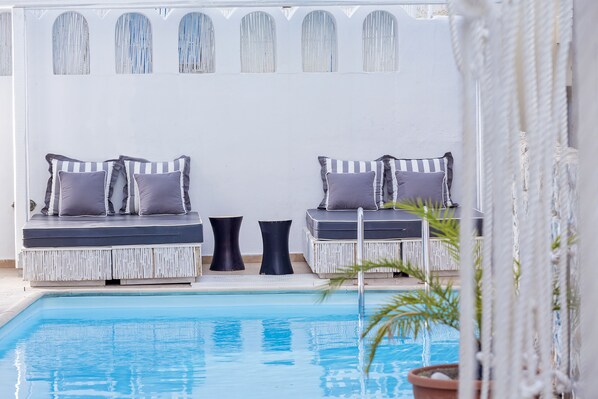 Un área de estar junto a la piscina con dos bancos acolchados y una mesita, rodeada por un muro blanco con ventanas de arco.
