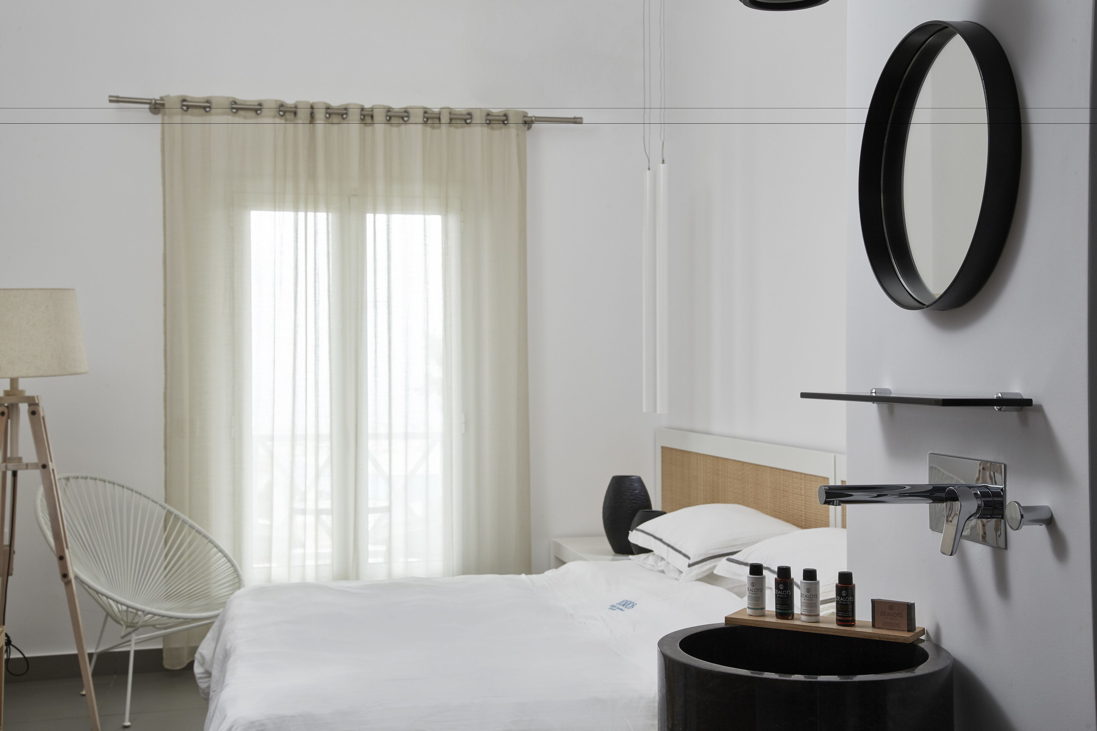 A hotel room with a bed, a bedside table, a lamp, a chair, a mirror, and a wall-mounted shelf with toiletries.