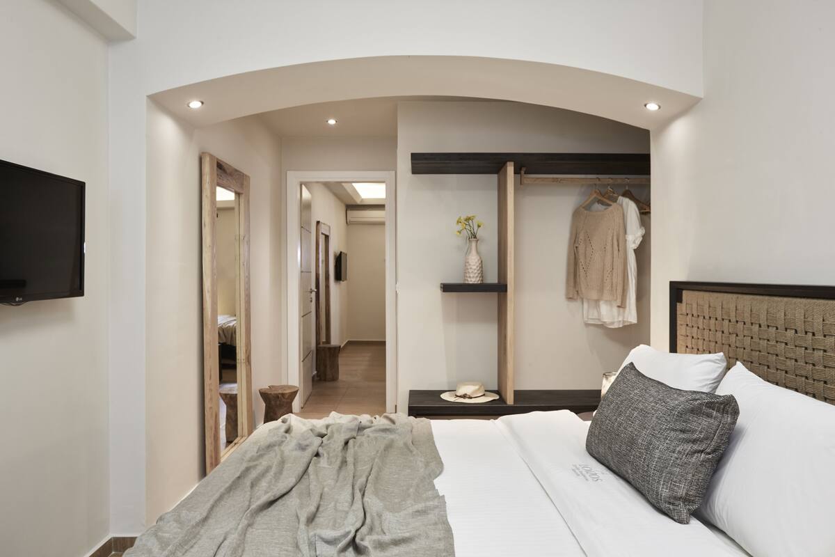 A modern hotel room with a large bed, a flat-screen TV, a mirror, a vanity with a sink, and a hallway leading to other rooms.
