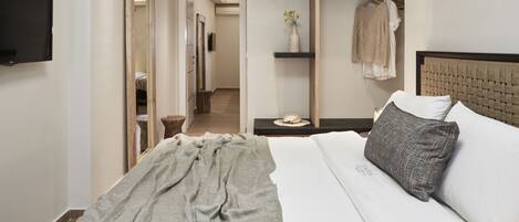 A modern hotel room with a large bed, a flat-screen TV, a mirror, a vanity with a sink, and a hallway leading to other rooms.