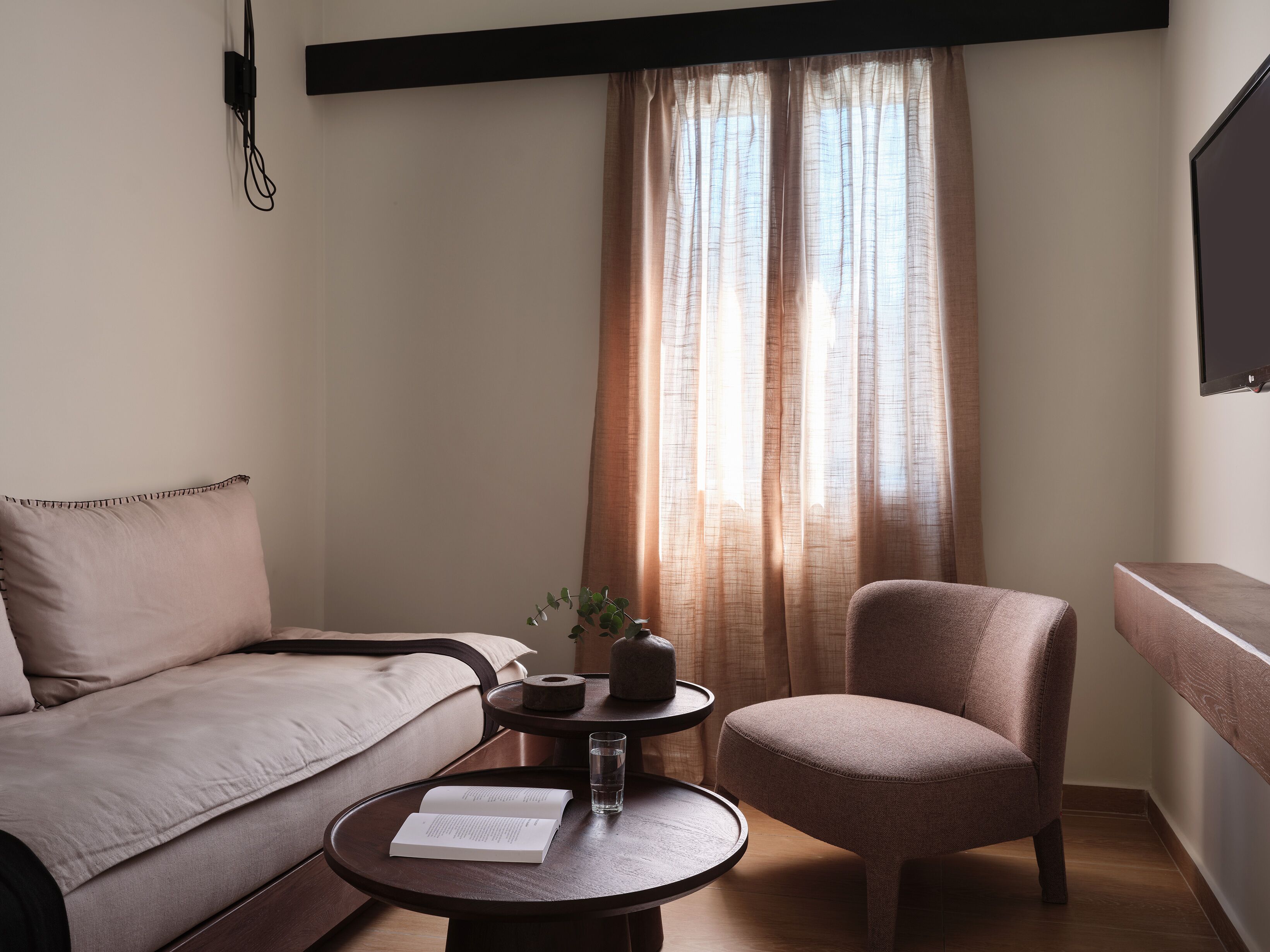 Una habitación acogedora con un sofá, una silla, una mesita con un libro y un vaso de agua, y una ventana con cortinas.