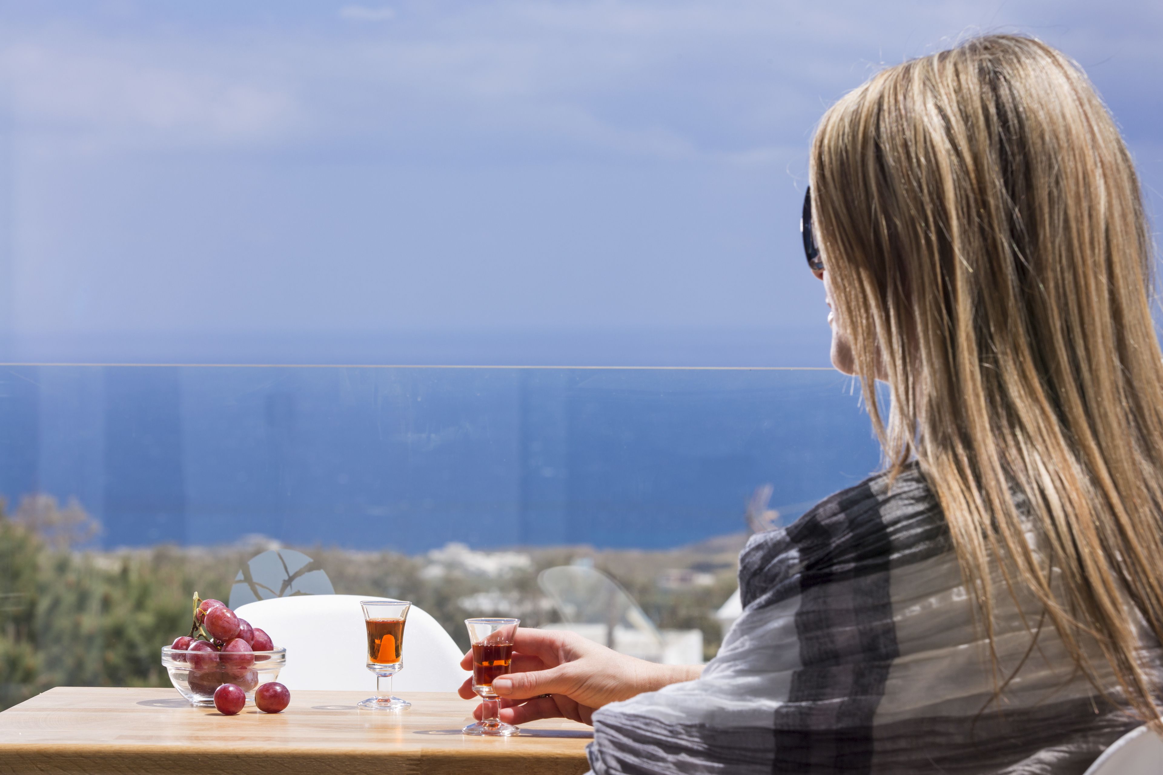 A person seated at a table with a view of the sea, a bowl of grapes, and two glasses of a dark beverage.