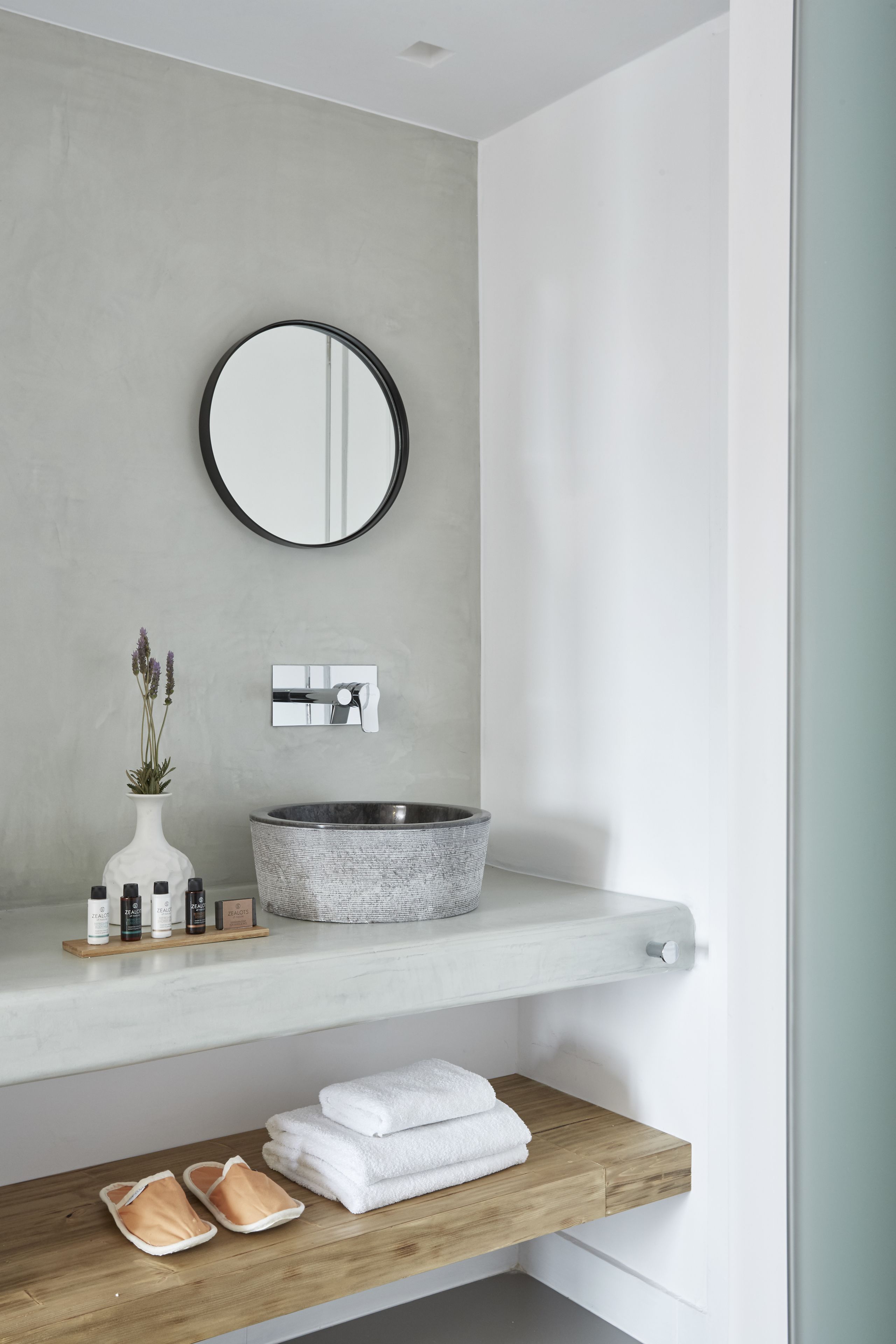 A bathroom with a round mirror, a round sink, a small vase with flowers, and a wooden shelf with towels and slippers.
