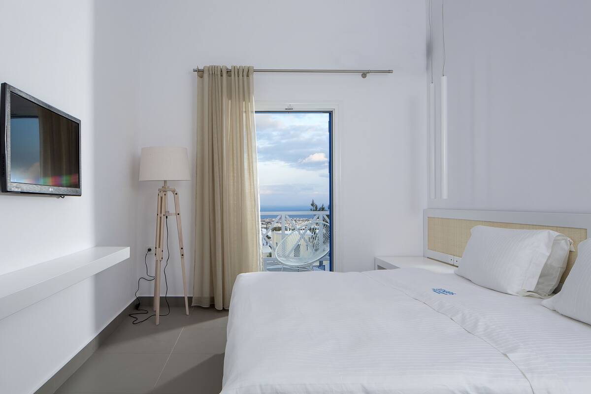 A hotel room with a bed, a TV, a lamp, and a view of the sea through a door.
