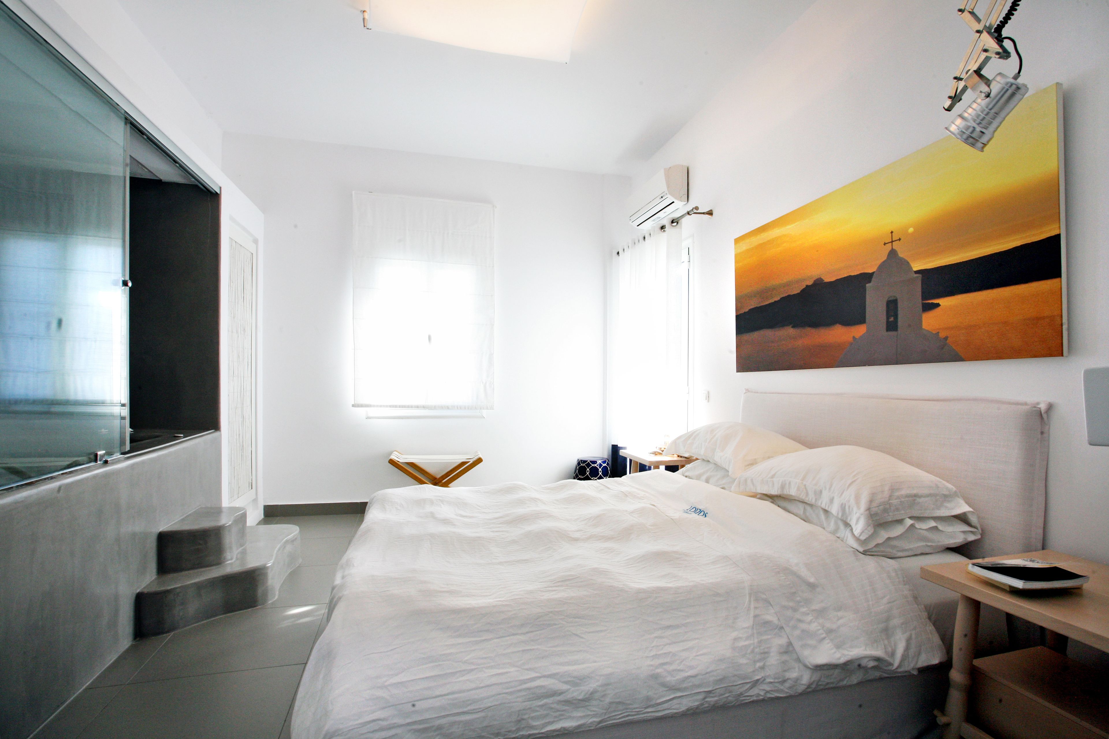 A bedroom with a bed, a nightstand, a window with curtains, and a painting of a sunset over a church.