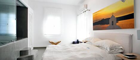 A bedroom with a bed, a nightstand, a window with curtains, and a painting of a sunset over a church.