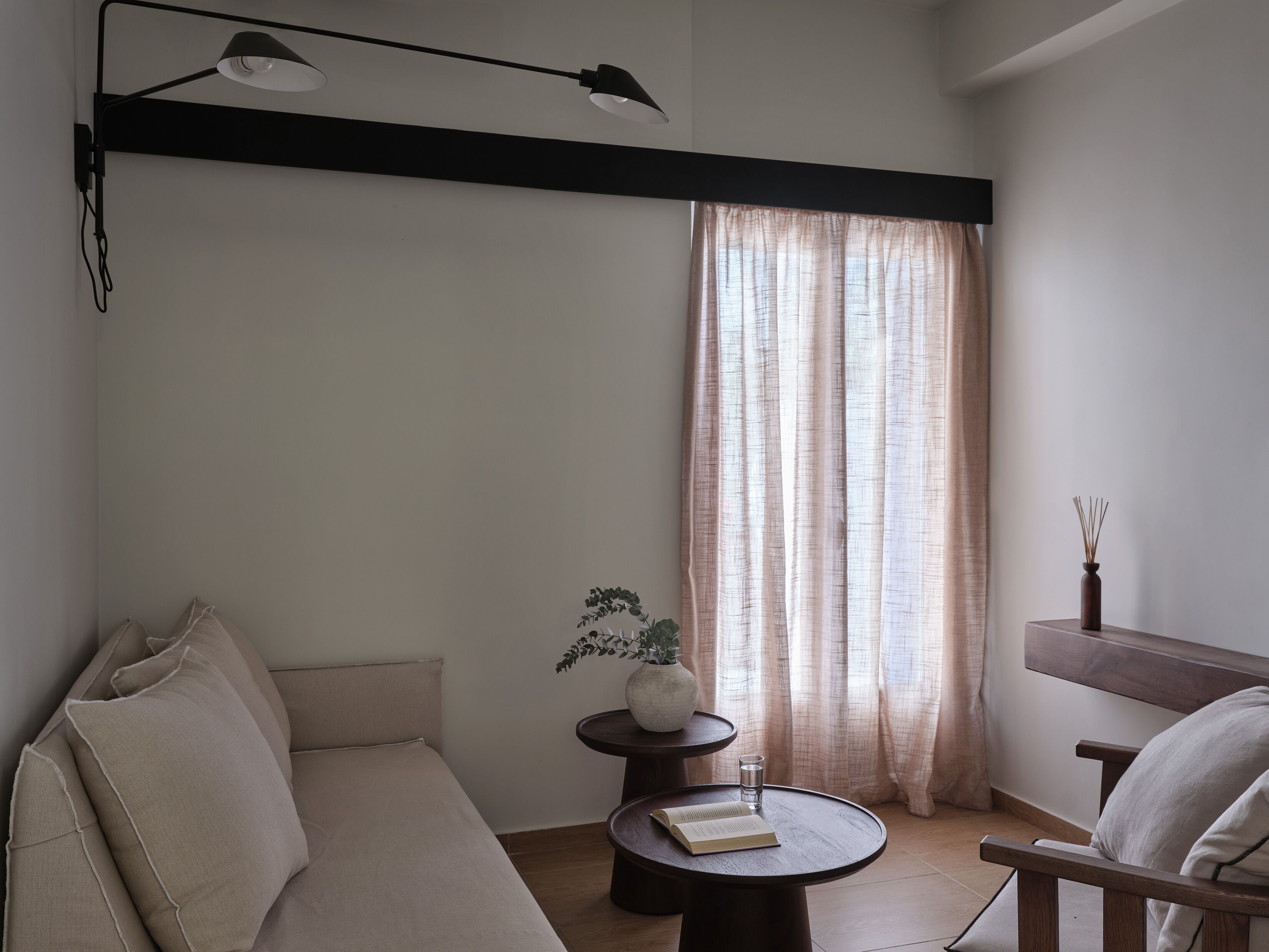 A minimalist living room with a sofa, a small round table with a book and glass, a potted plant, and a window with sheer curtains.