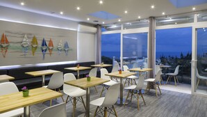 A modern dining area with tables and chairs, a mural of sailboats, and large windows offering a view of the city. - Loizos Stylish Residences (Santorini)