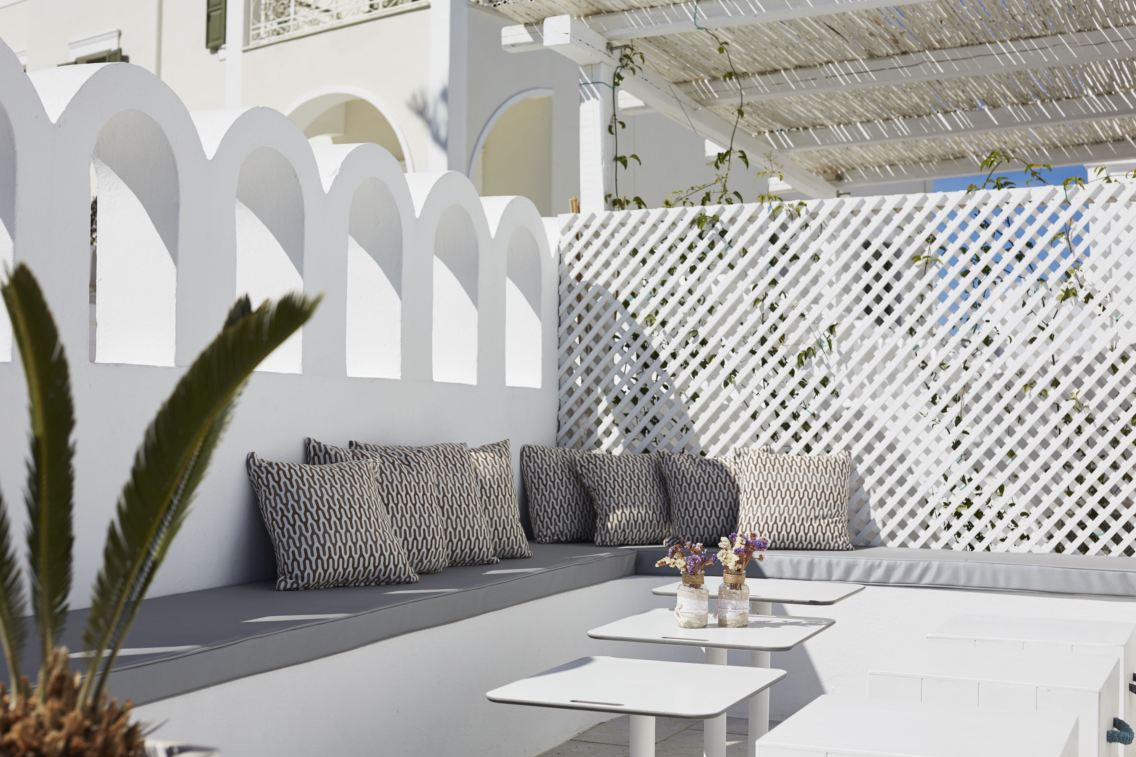 Un espacio al aire libre moderno con mobiliario blanco y cojines estampados, rodeado por una cerca de celosía blanca y un edificio blanco al fondo.